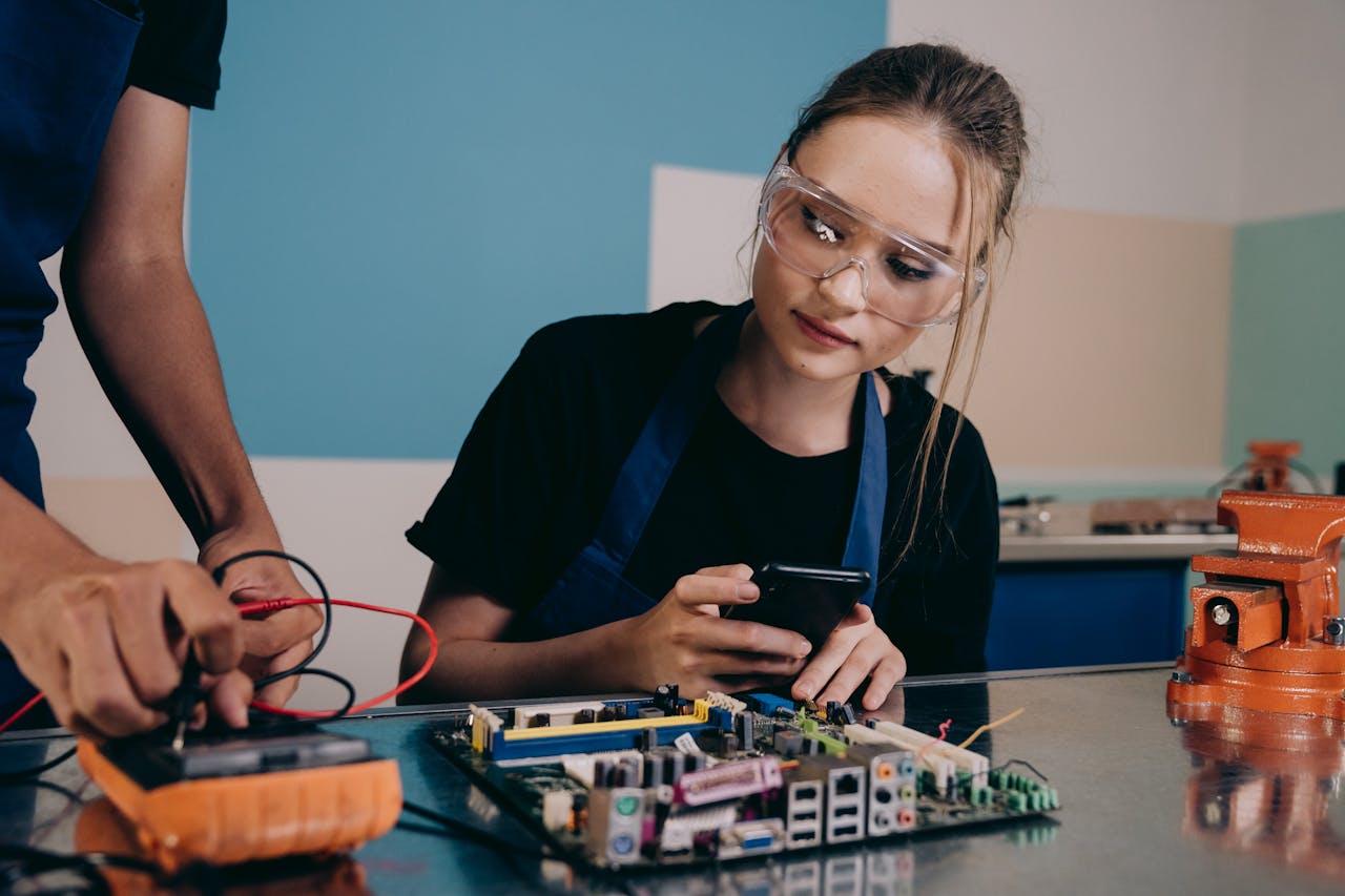 Services Female technician examining a circuit board in an electronics lab, using a smartphone for reference.